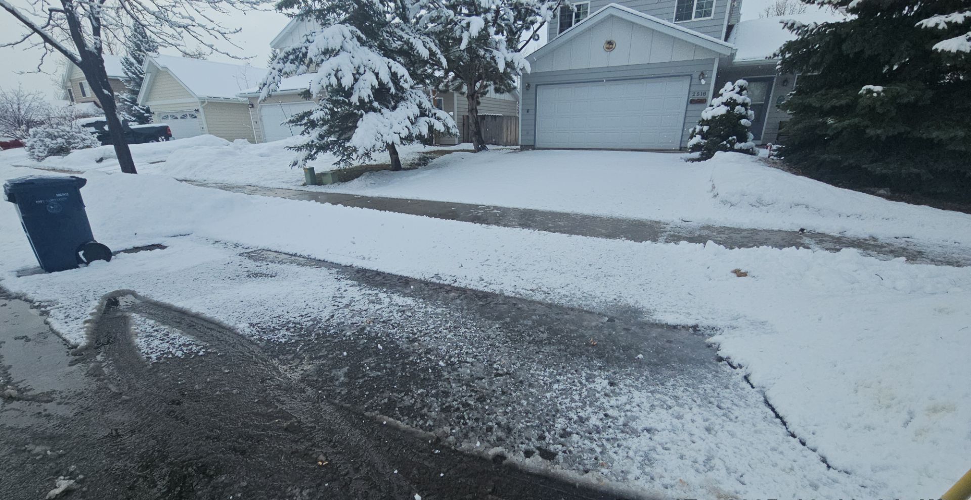 A suburban street covered in fresh snow with a house, a garage, and a trash bin in the foreground.