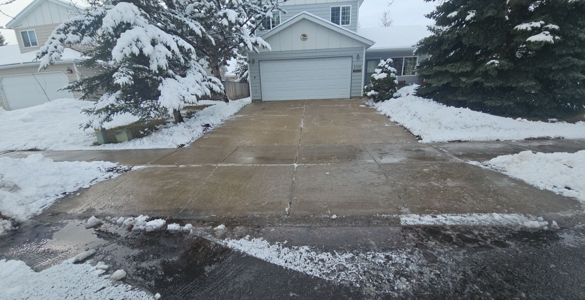 A residential driveway and walkway are covered in melting snow, leading to a house with a white garage door.