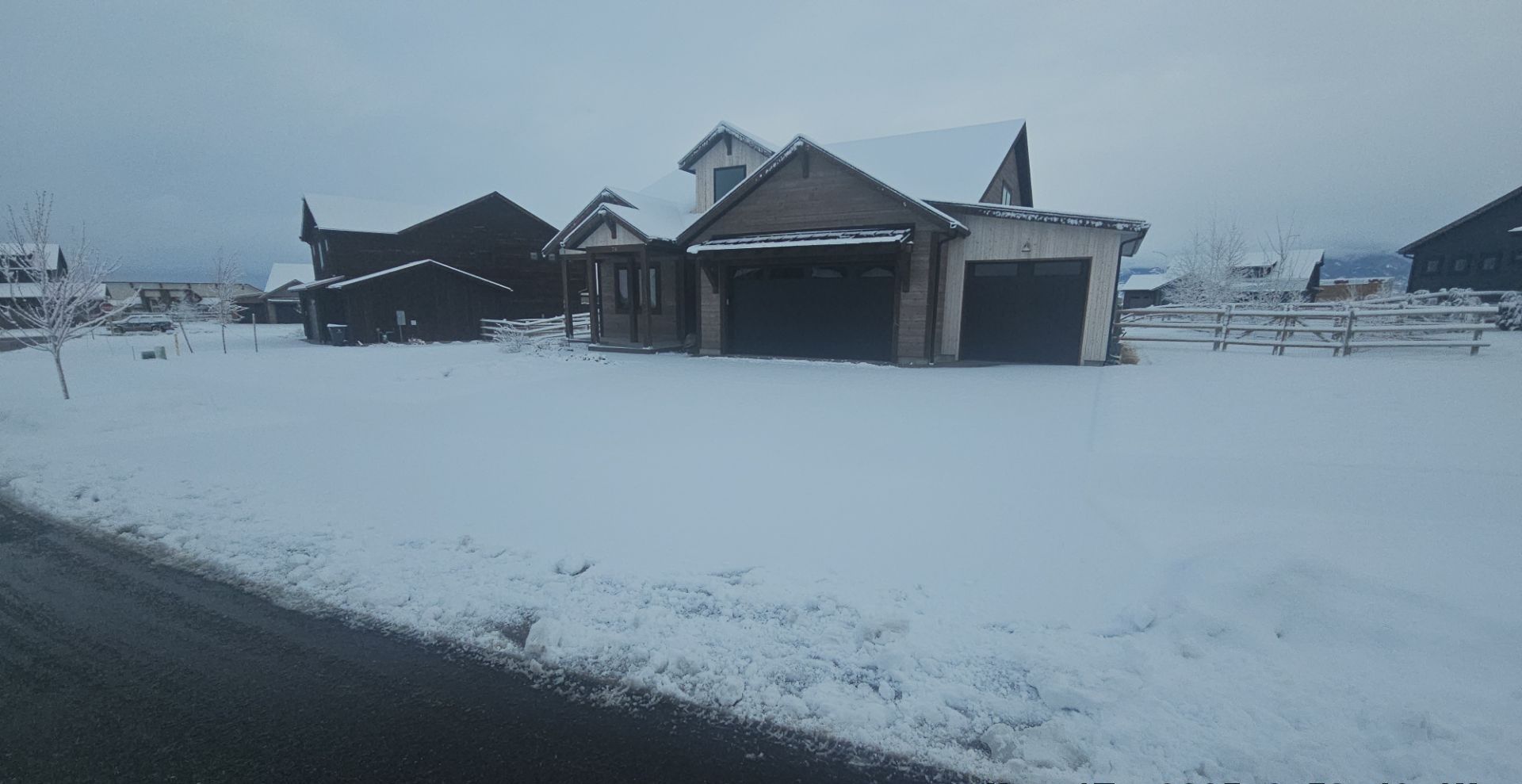 A snowy residential landscape featuring a dark house with a two-car garage under an overcast winter sky.