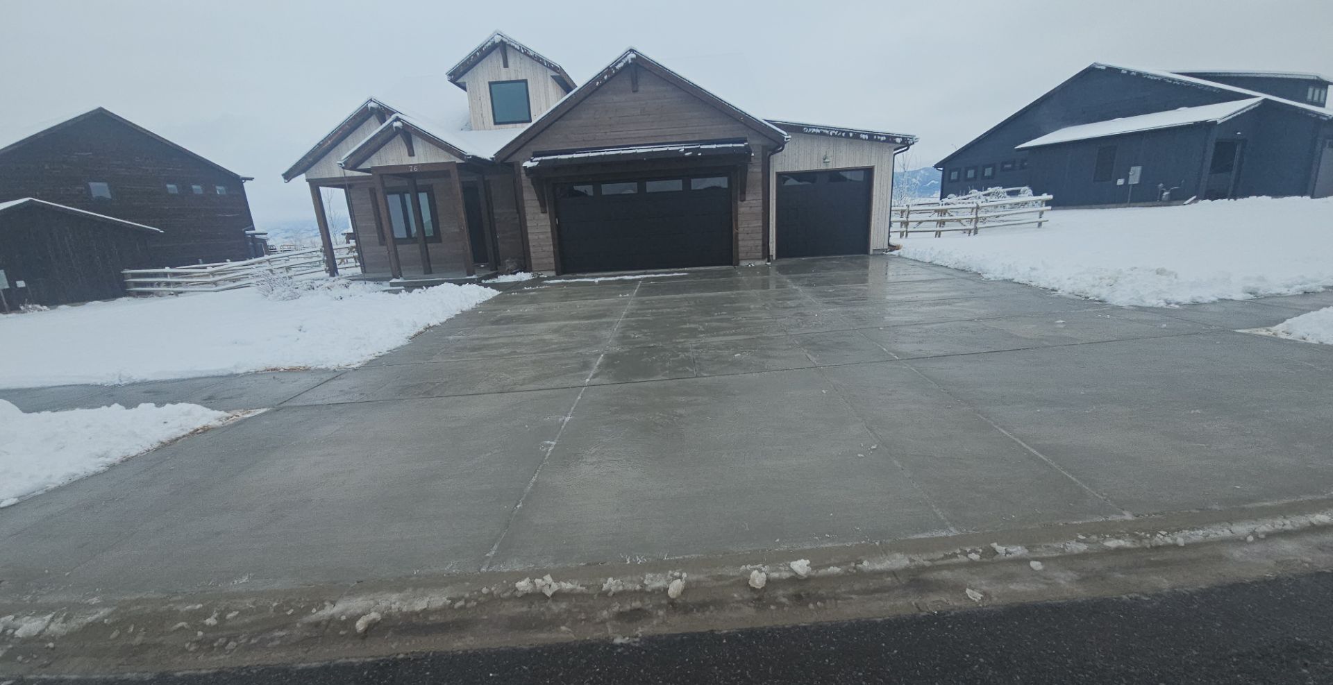 A modern, single-story house with a garage and a wet concrete driveway during a light snowfall in a residential area.