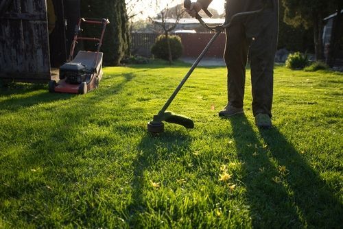 A person trims grass with a string trimmer in a sunlit yard, with a lawnmower parked in the background.