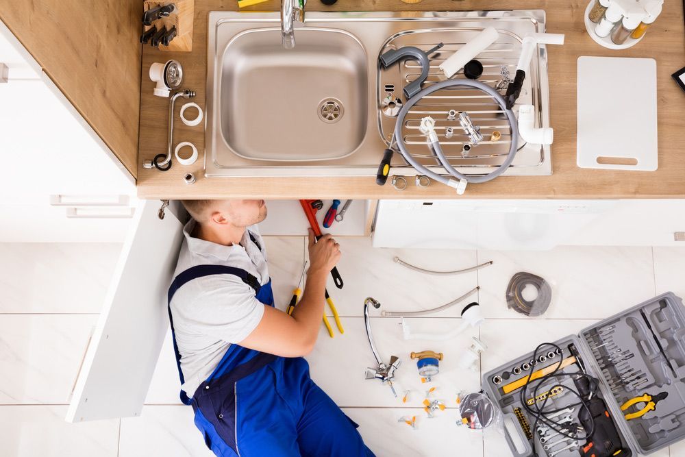 Plumber in blue overalls working on a kitchen sink, surrounded by tools.