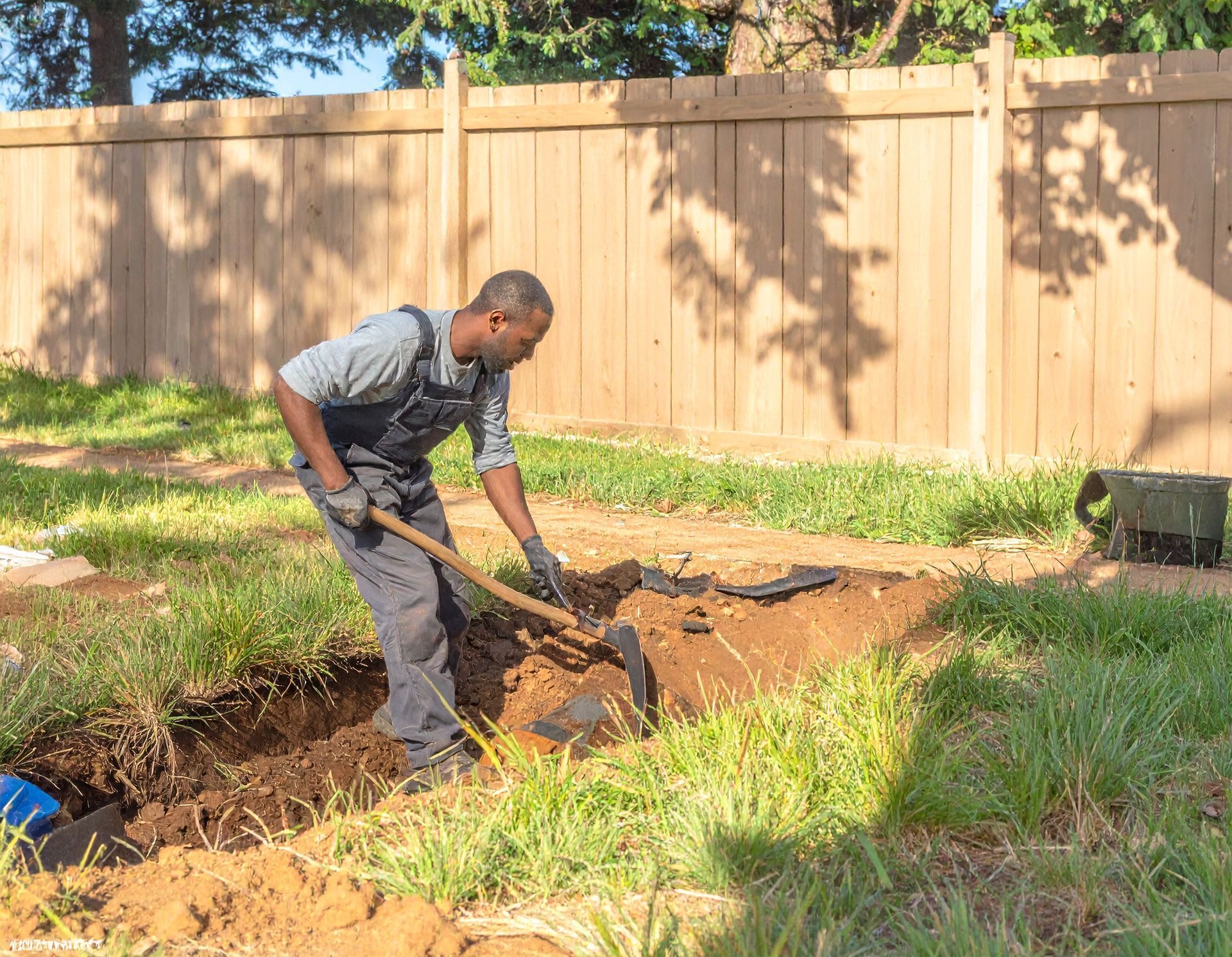 Person digging a trench in a grassy backyard beside a wooden fence