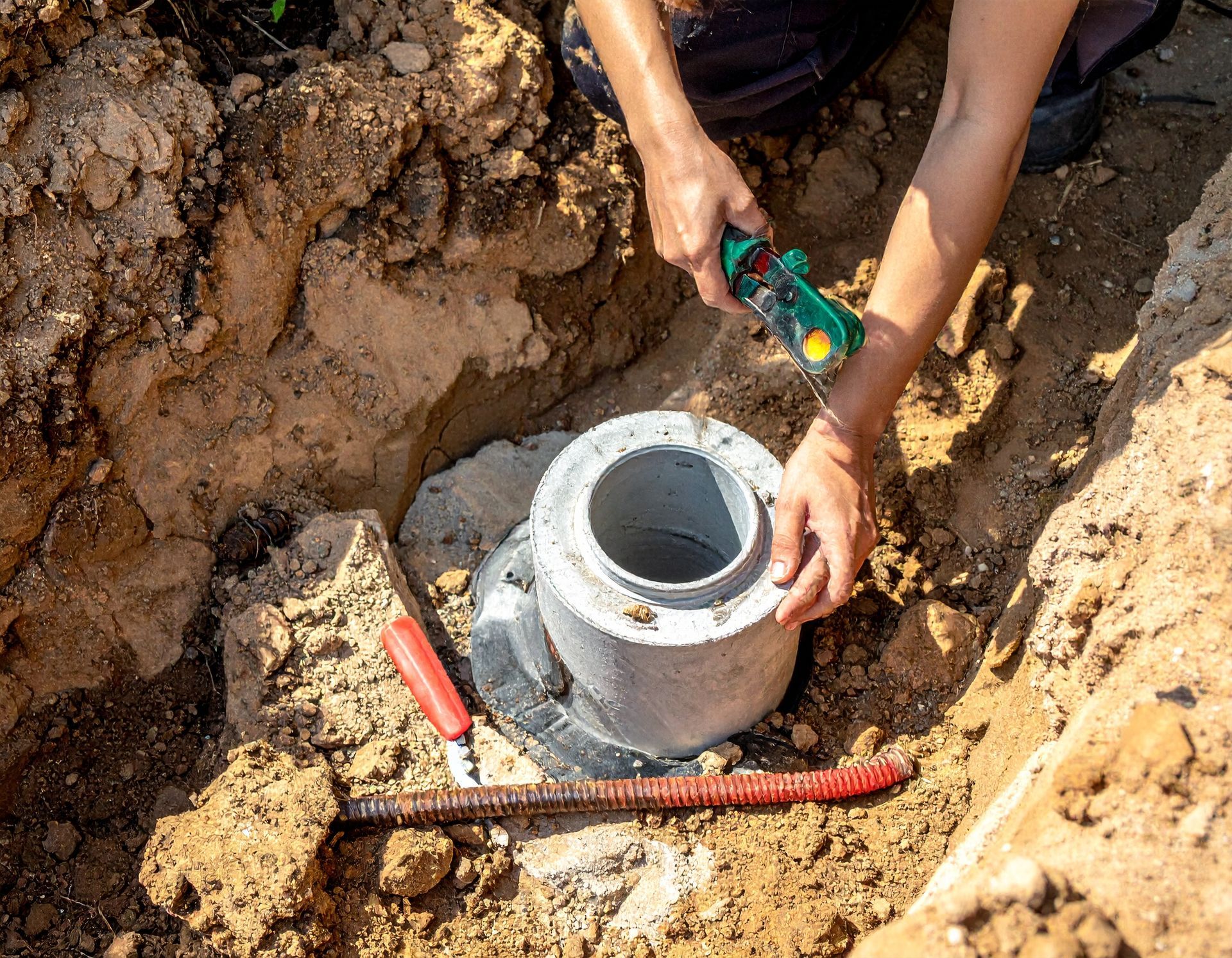 Hands using tools to install a gray PVC pipe in a dirt excavation site