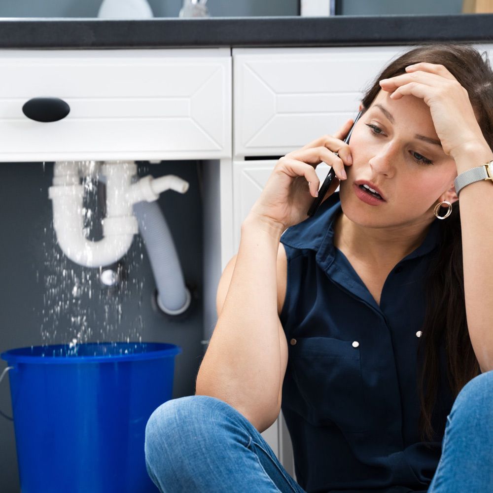 Woman with a concerned expression on the phone next to a leaking sink, a blue bucket catching water.