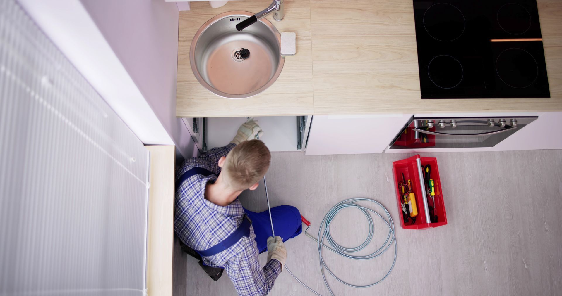 Plumber working under a kitchen sink. Tools are on the floor. White and light wood tones.