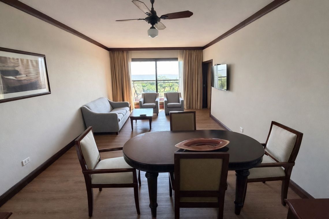 Comedor con mesa redonda, cuatro sillas y un sofá junto a una ventana en una luminosa habitación de hotel.