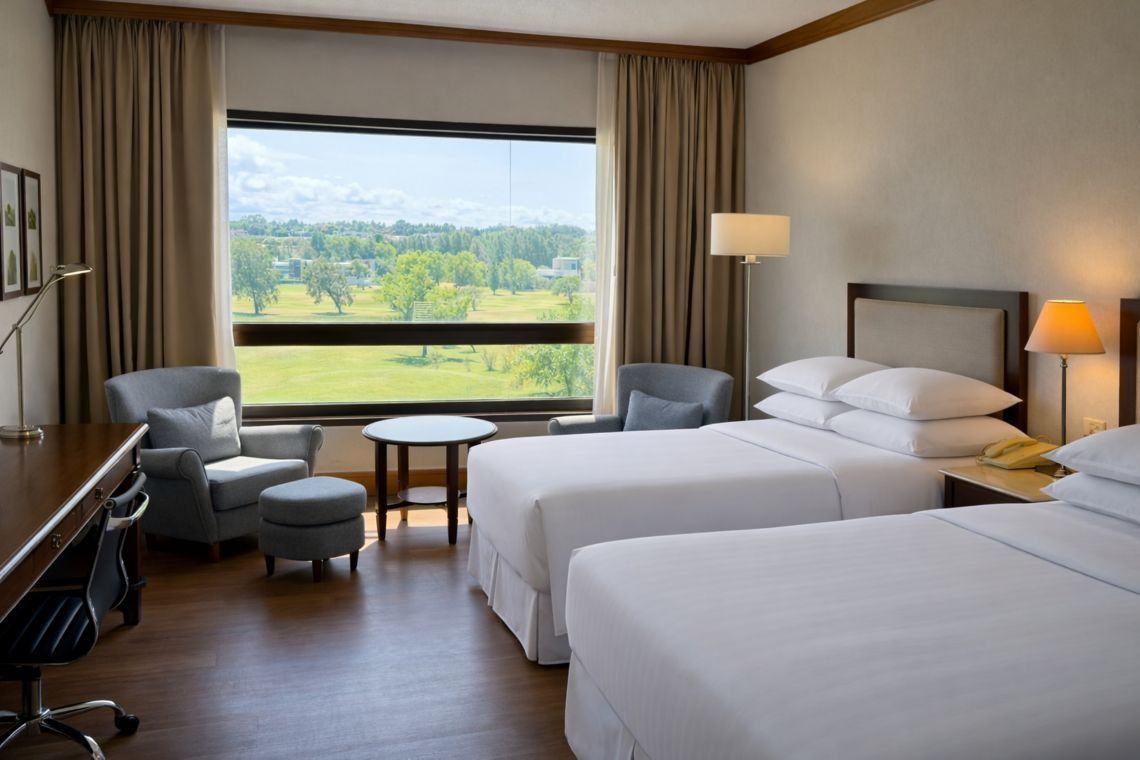 Habitación de hotel con dos camas, ventana grande, sillón y lámparas de noche cálidas con vistas a campos verdes.