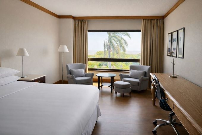 Habitación de hotel con dos camas, zona de estar, escritorio y una gran ventana con vistas a las palmeras.
