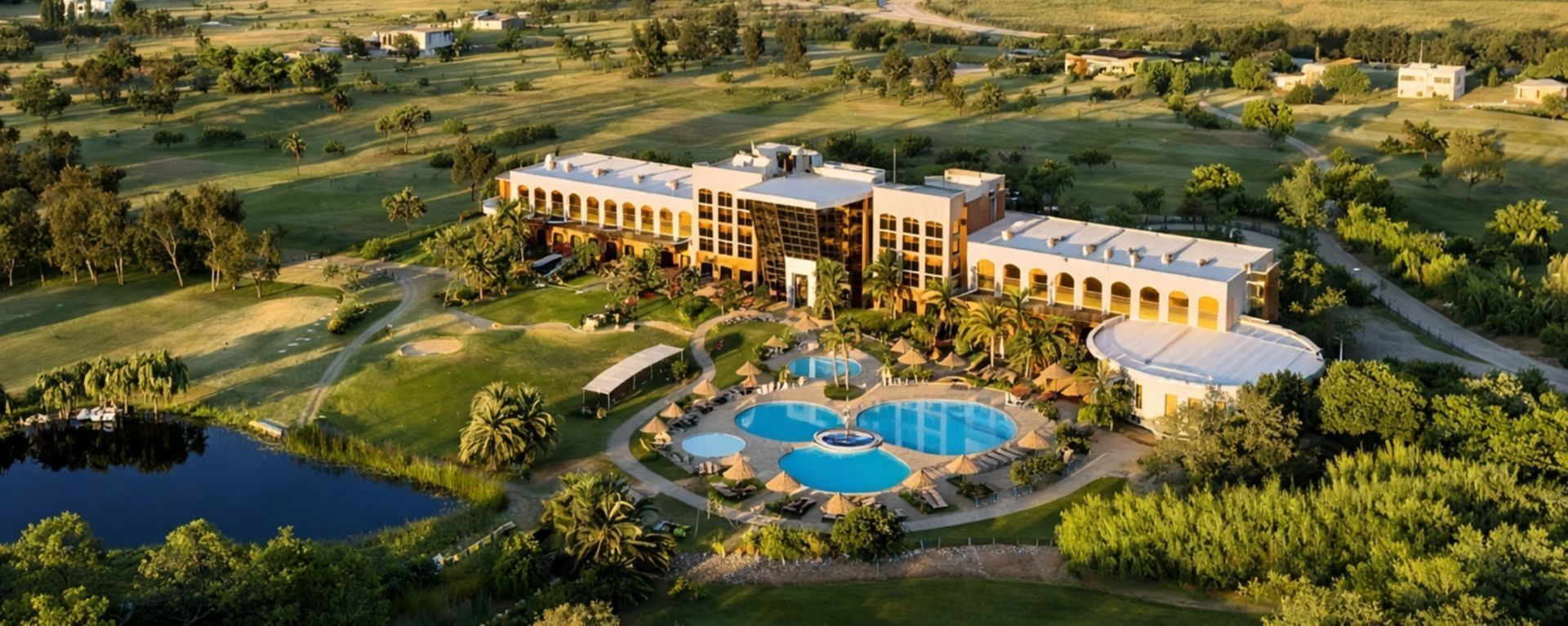 Vista aérea de un complejo turístico con una gran piscina, vegetación circundante y jardines paisajísticos al atardecer.