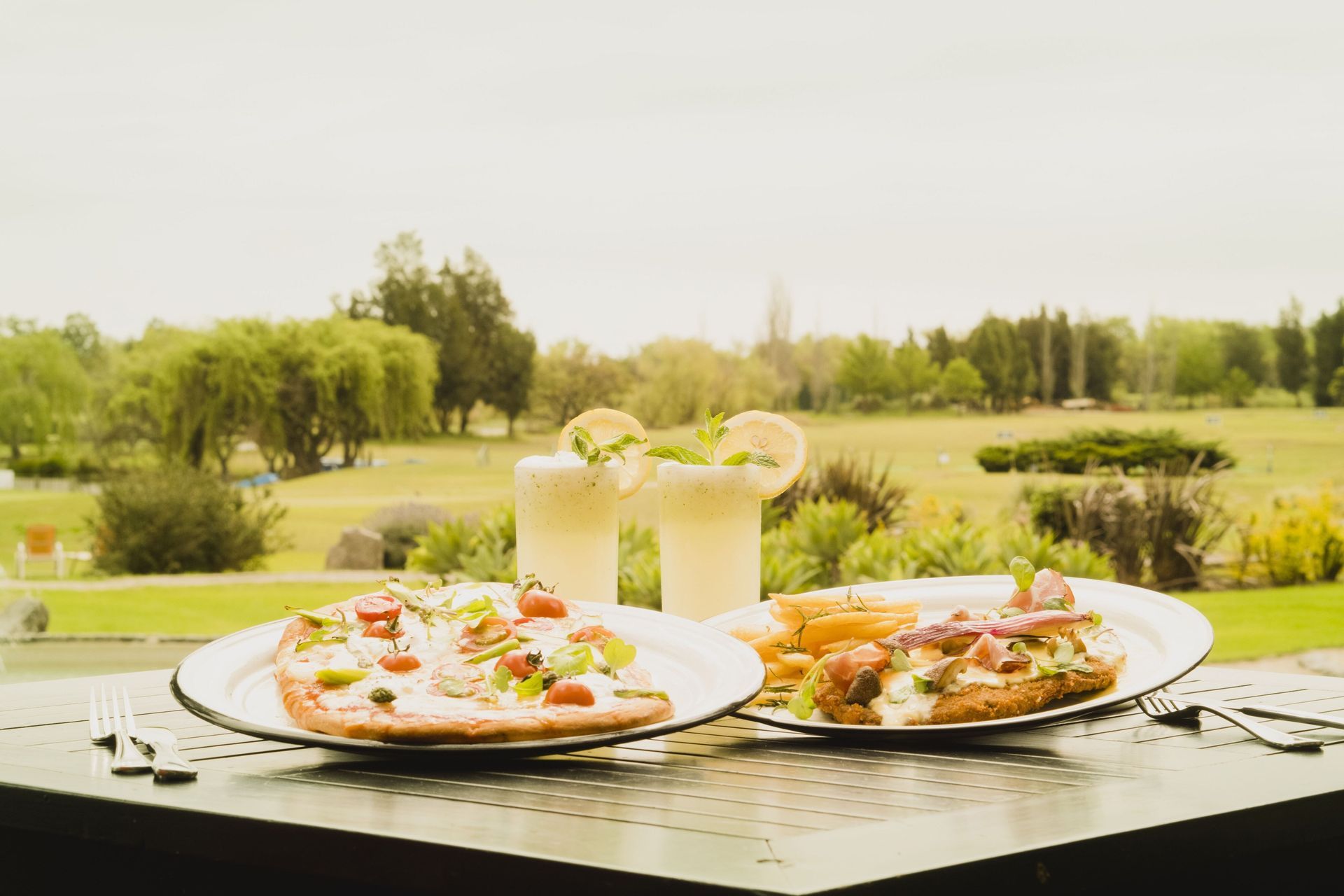 Mesa al aire libre con comida servida en plato y dos bebidas, enmarcada por un paisaje verde y soleado.