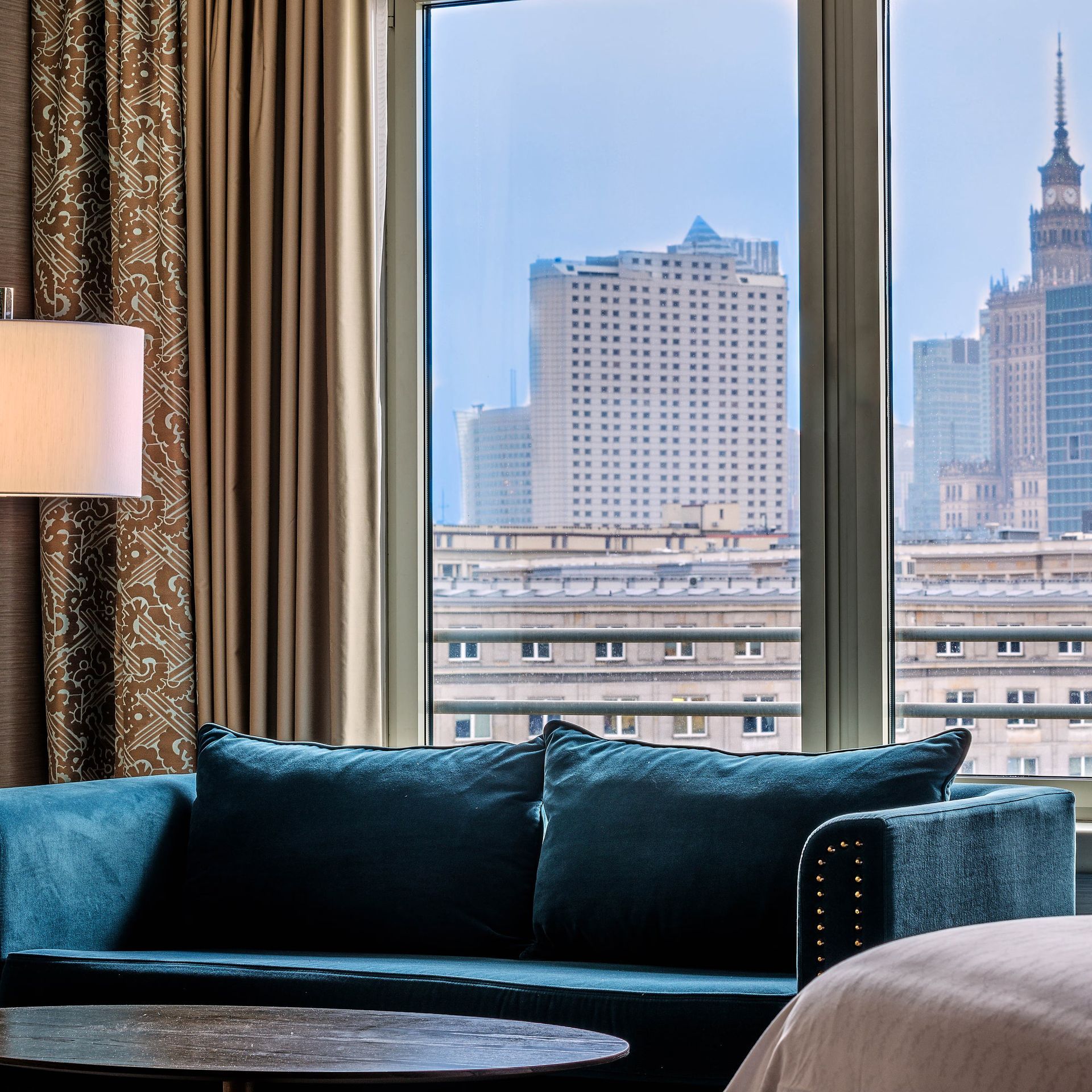 Sofá color turquesa junto a una ventana con vistas a los rascacielos de la ciudad en una habitación de hotel moderna.