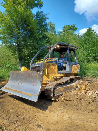 Yellow bulldozer on a dirt lot beneath trees and blue sky
