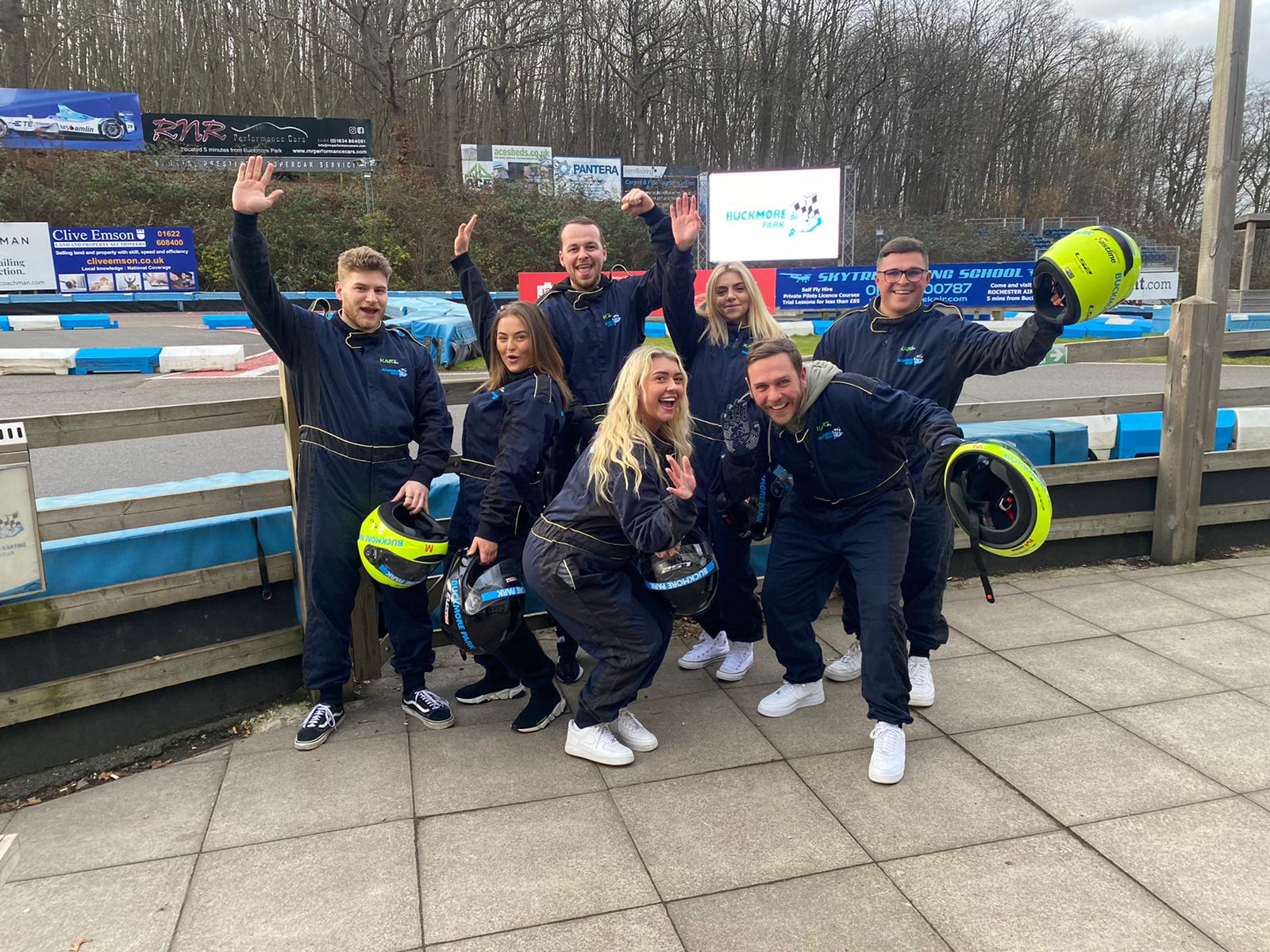 Group of six posing and cheering beside a race car, with a blue barrier and snowy track in the background.