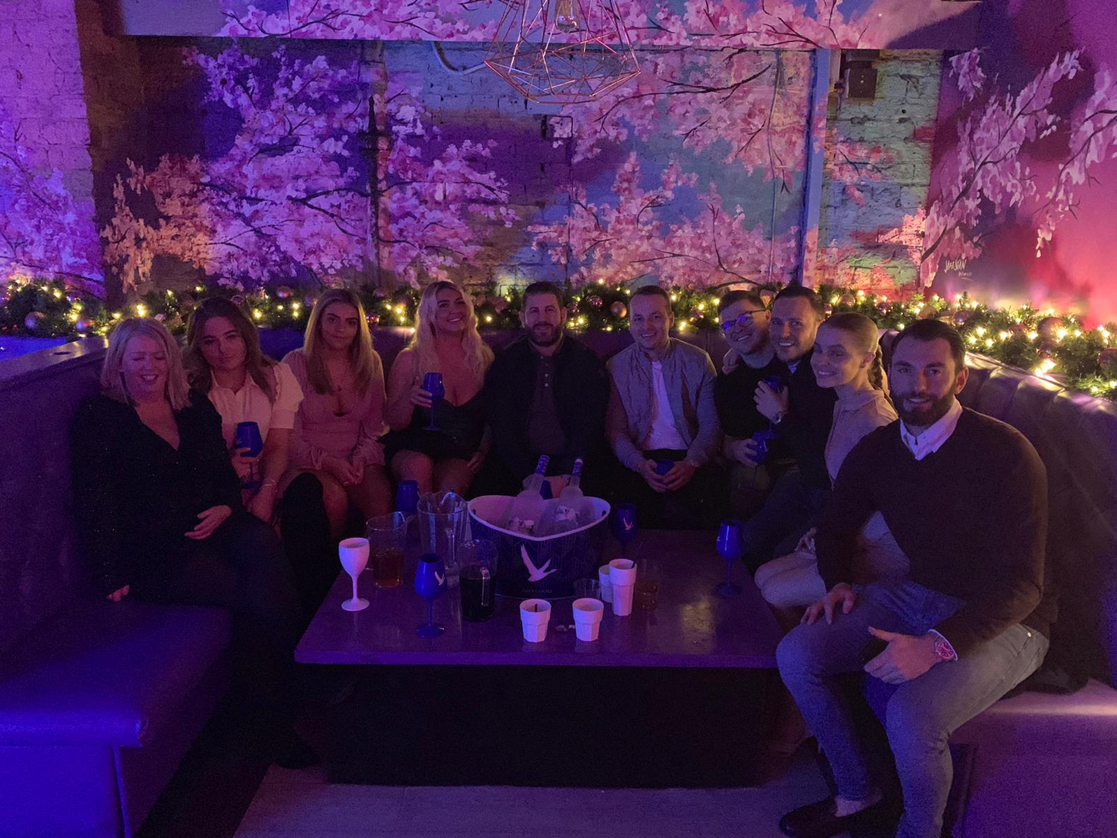 Group seated around a table in a purple-lit lounge with string lights and drinks