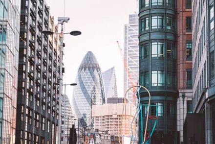 City street framed by tall buildings, with the Gherkin skyscraper in the center under a cloudy sky