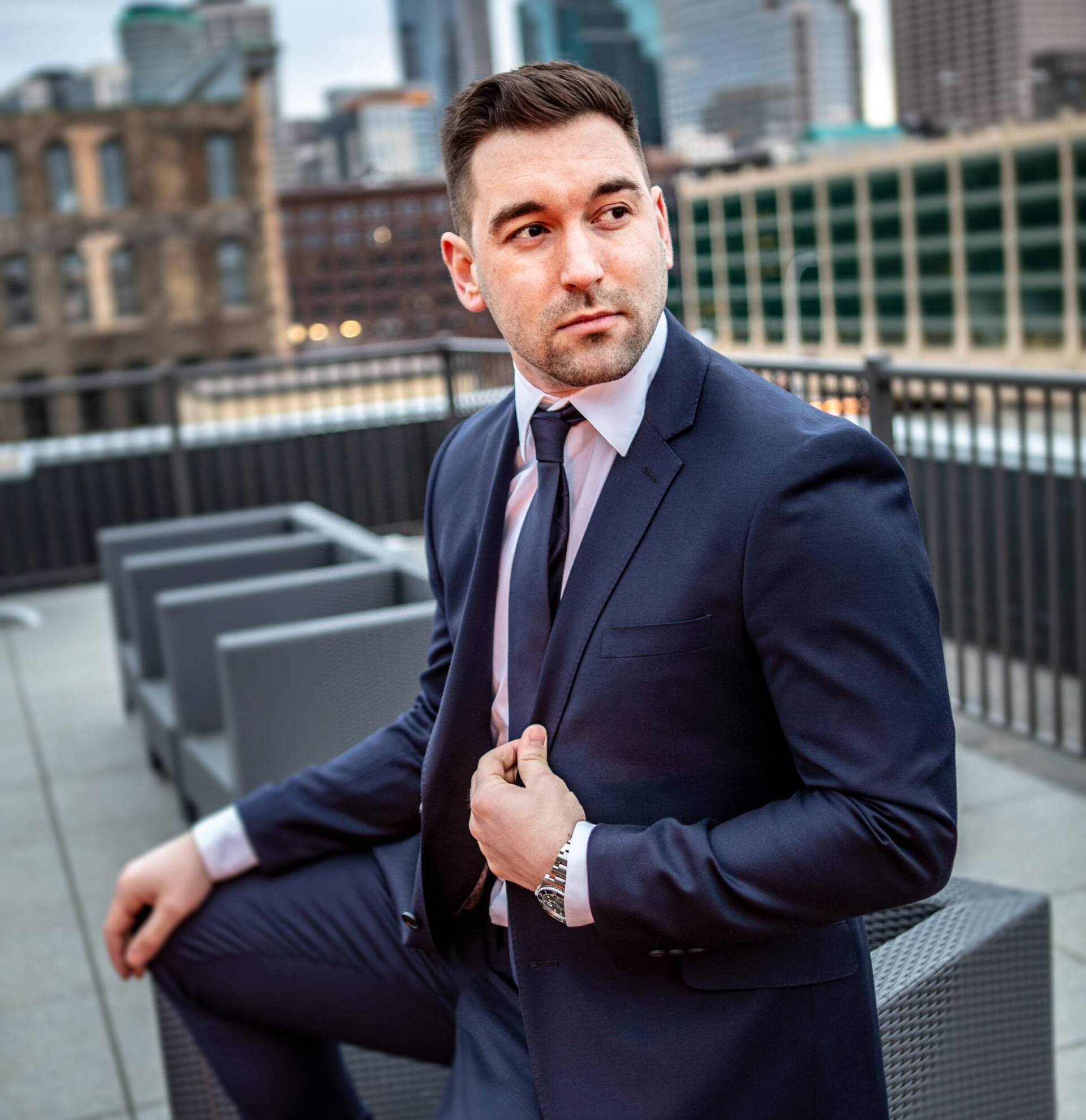 Man in a navy suit sitting on a rooftop terrace, looking to the side with city buildings behind him