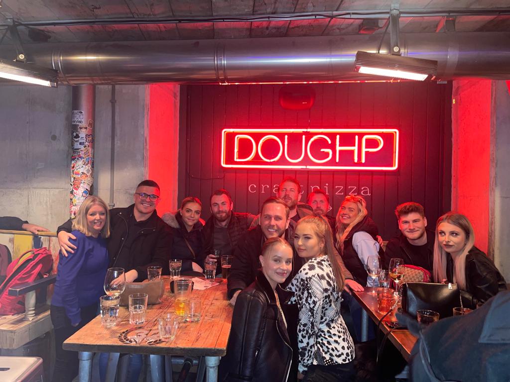 Group of people seated at tables in a neon-lit restaurant under a red “DOUGHP” sign.
