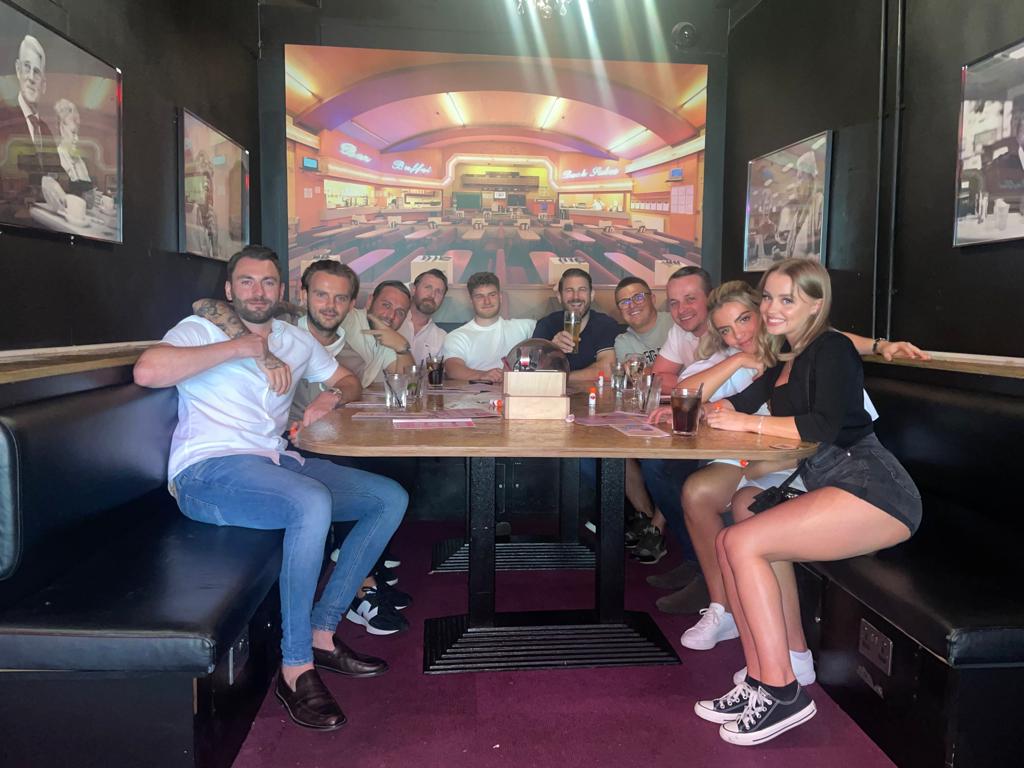 Group of people seated in a dimly lit restaurant booth, smiling around a table with drinks.