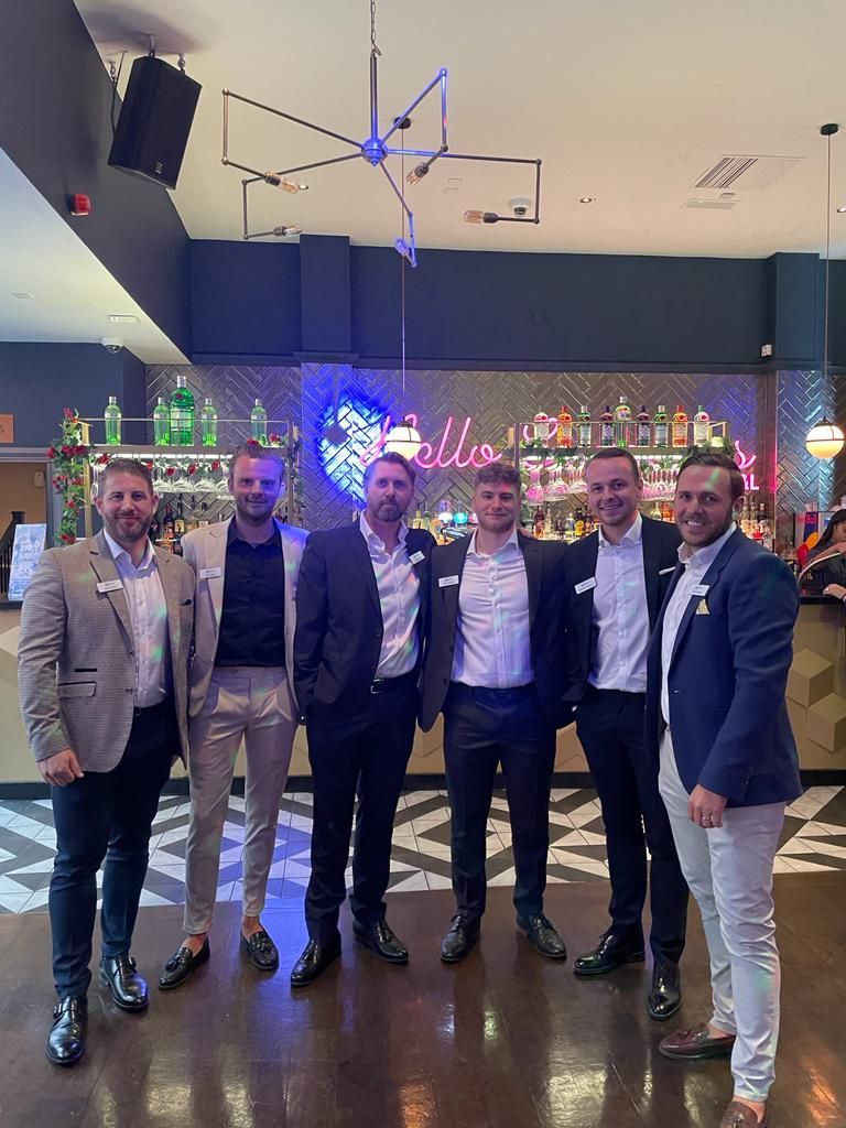 Six men in suits posing together at a bar with neon lights and a holiday wreath in the background