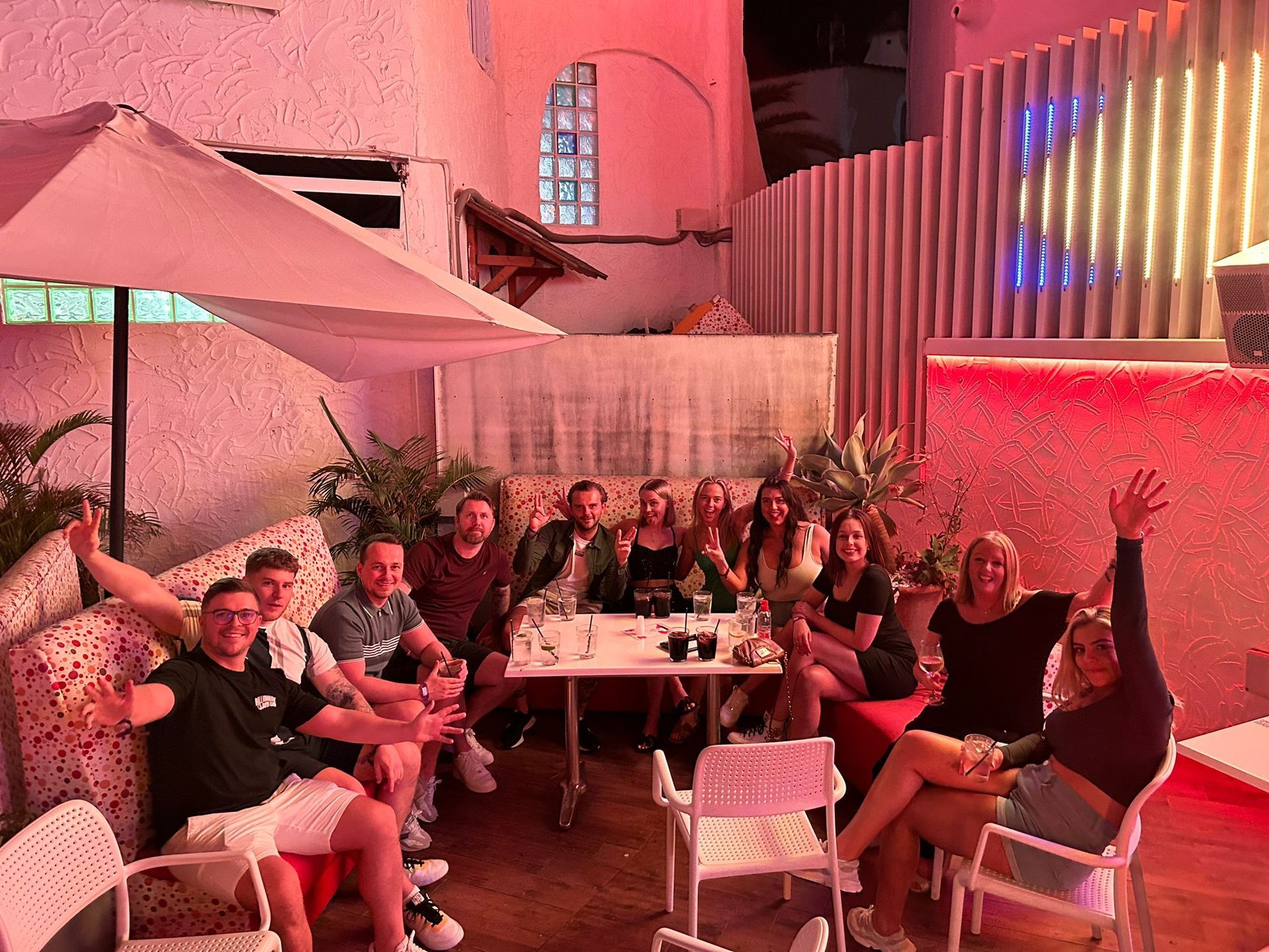 Group of people sitting and chatting outdoors under pink lighting at a patio table with umbrellas and plants