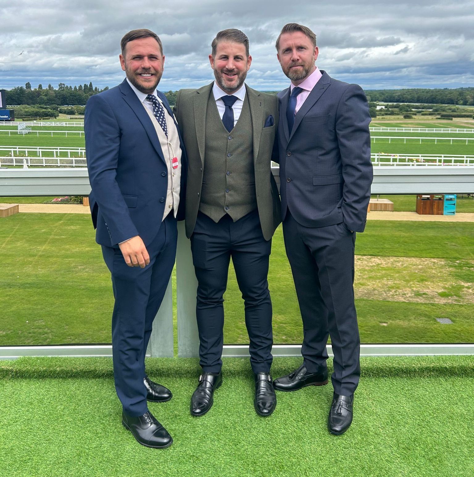 Three men in suits standing by a racetrack under cloudy skies