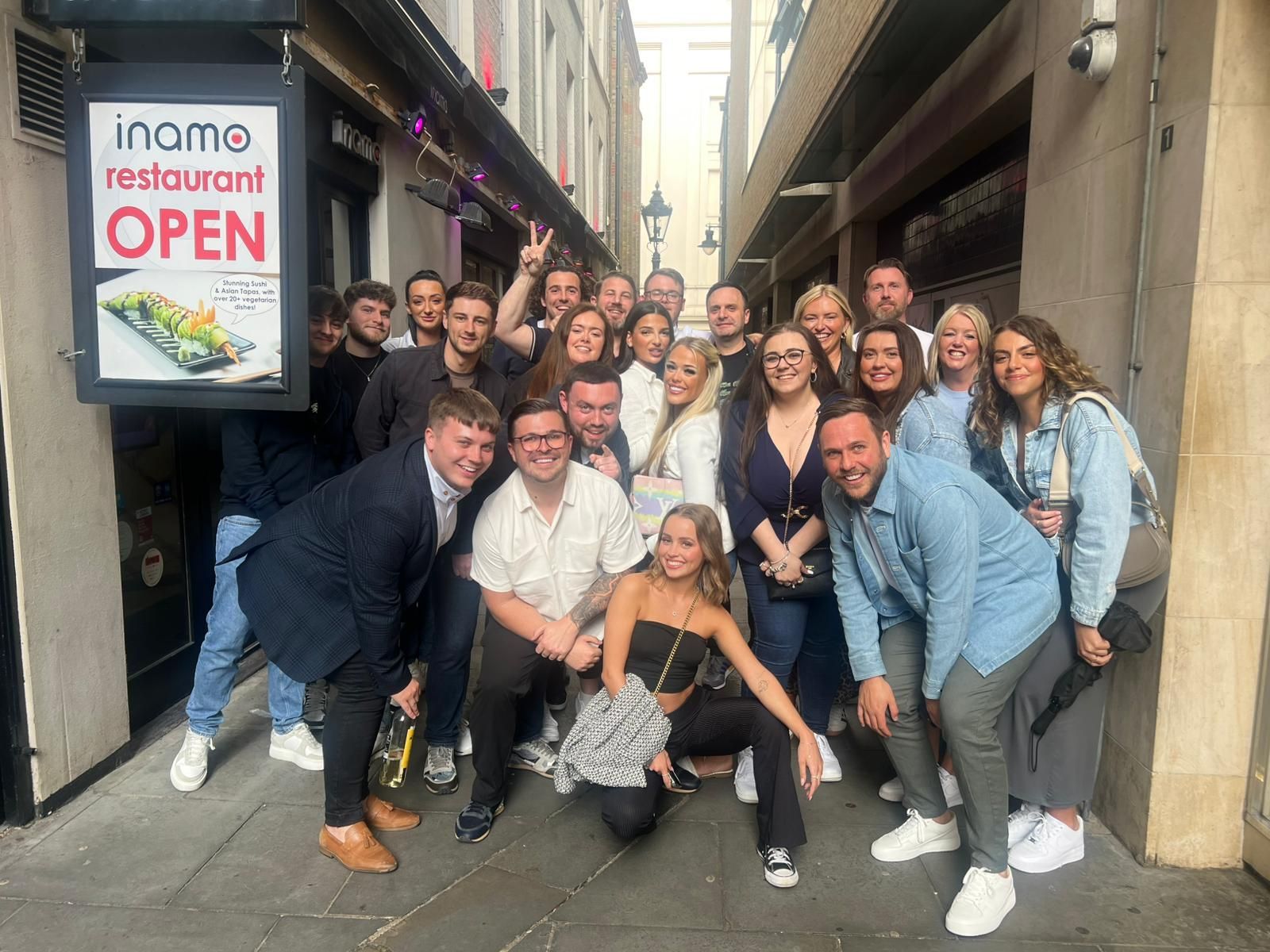 Group of people posing outside a restaurant with an “OPEN” sign on a narrow street.