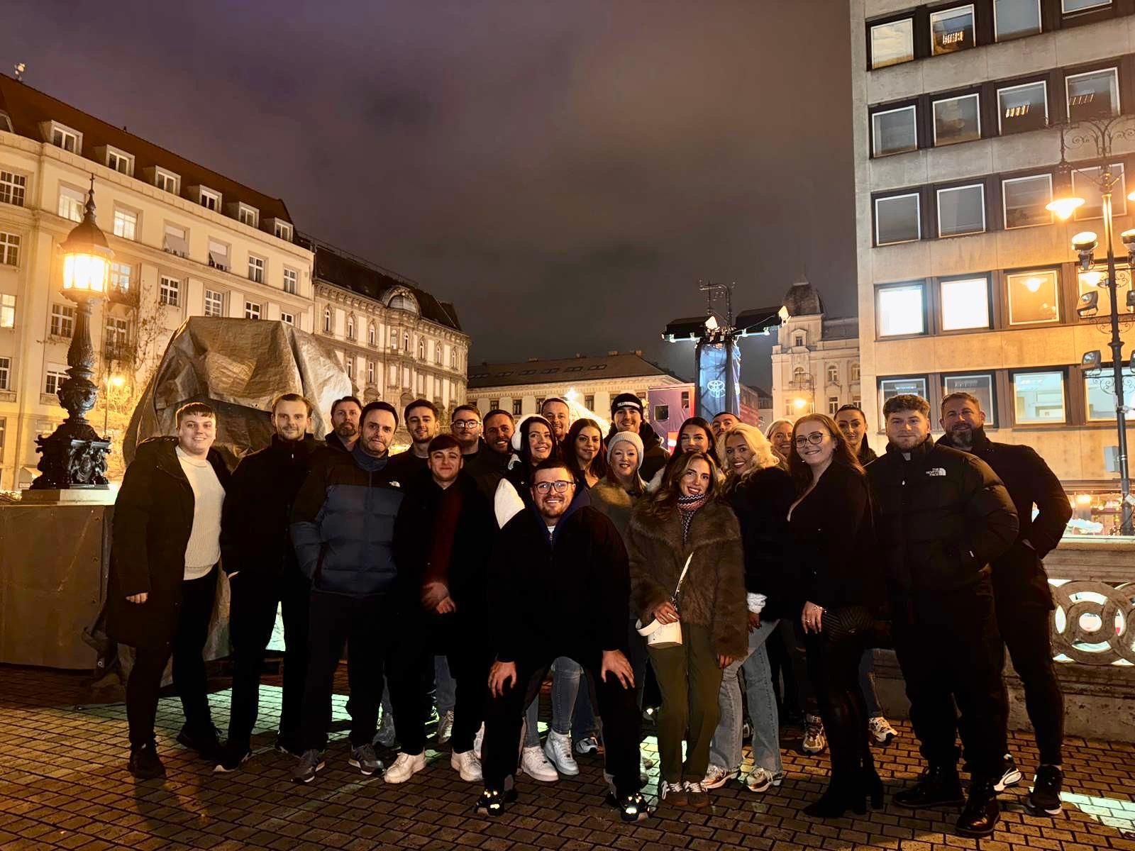 Group of people posing outdoors at night in a city square with lit buildings and a monument behind them
