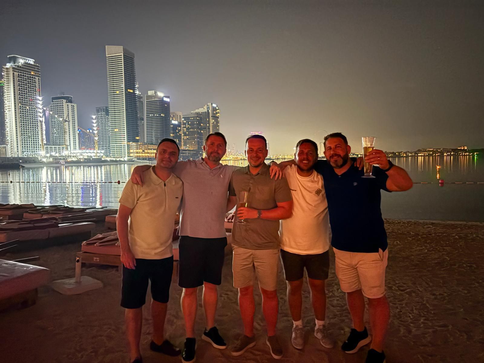 Five friends posing on a waterfront at night with city lights reflecting on the water
