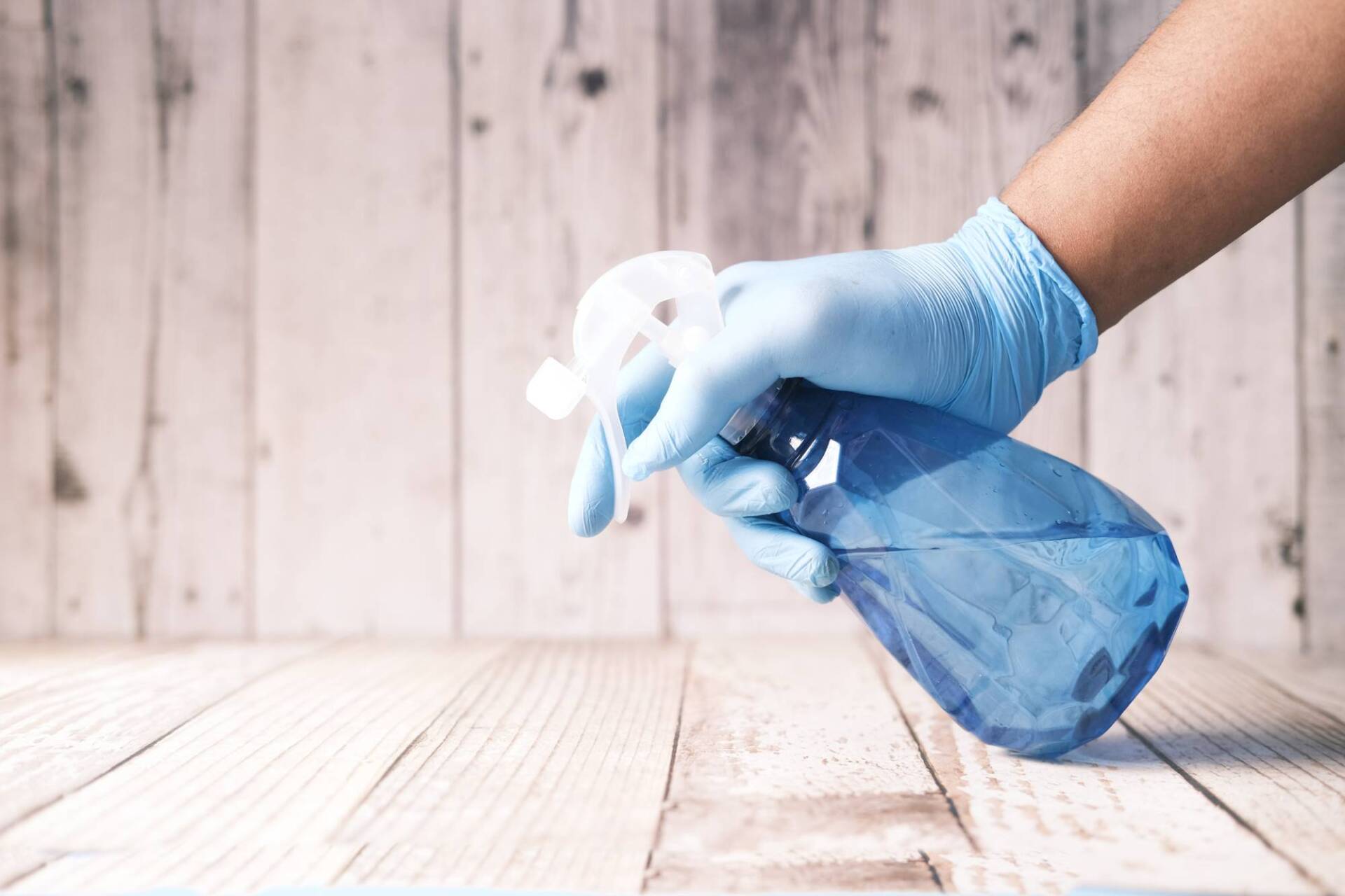 Gloved hand spraying cleaner on a wooden surface with a blue spray bottle