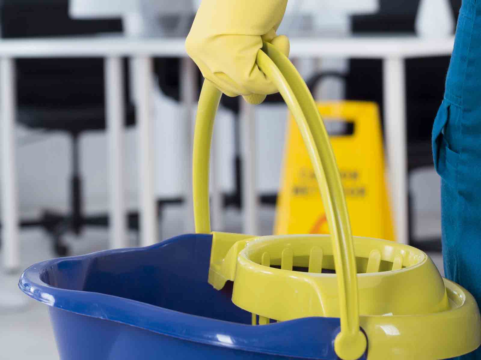 Yellow-gloved hand lifting a yellow bucket from a blue mop bucket in a cleaning area