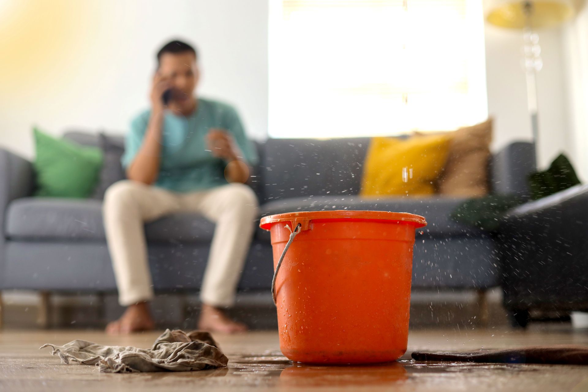 Orange bucket catching water leak on wet floor, with a blurred person sitting on a couch in the background