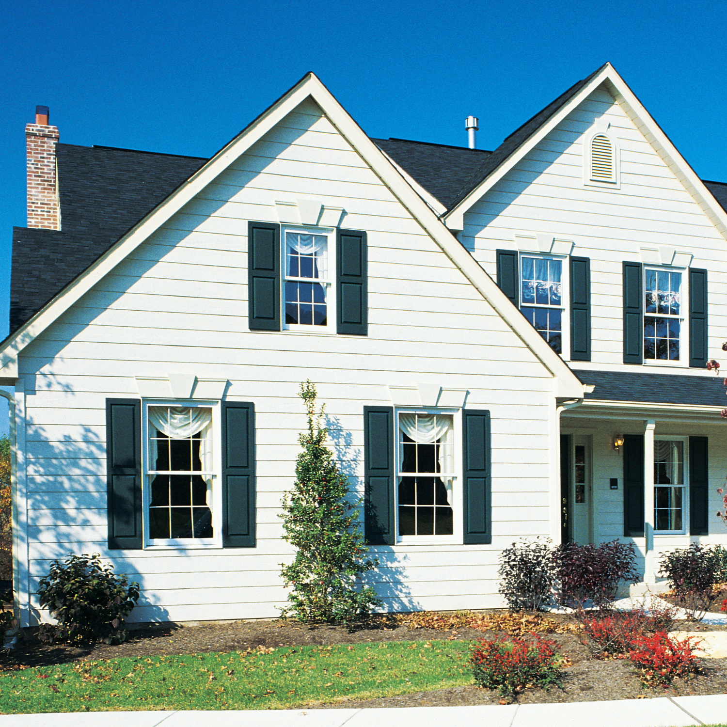 White suburban house with dark green shutters, front porch, and landscaped yard under a blue sky