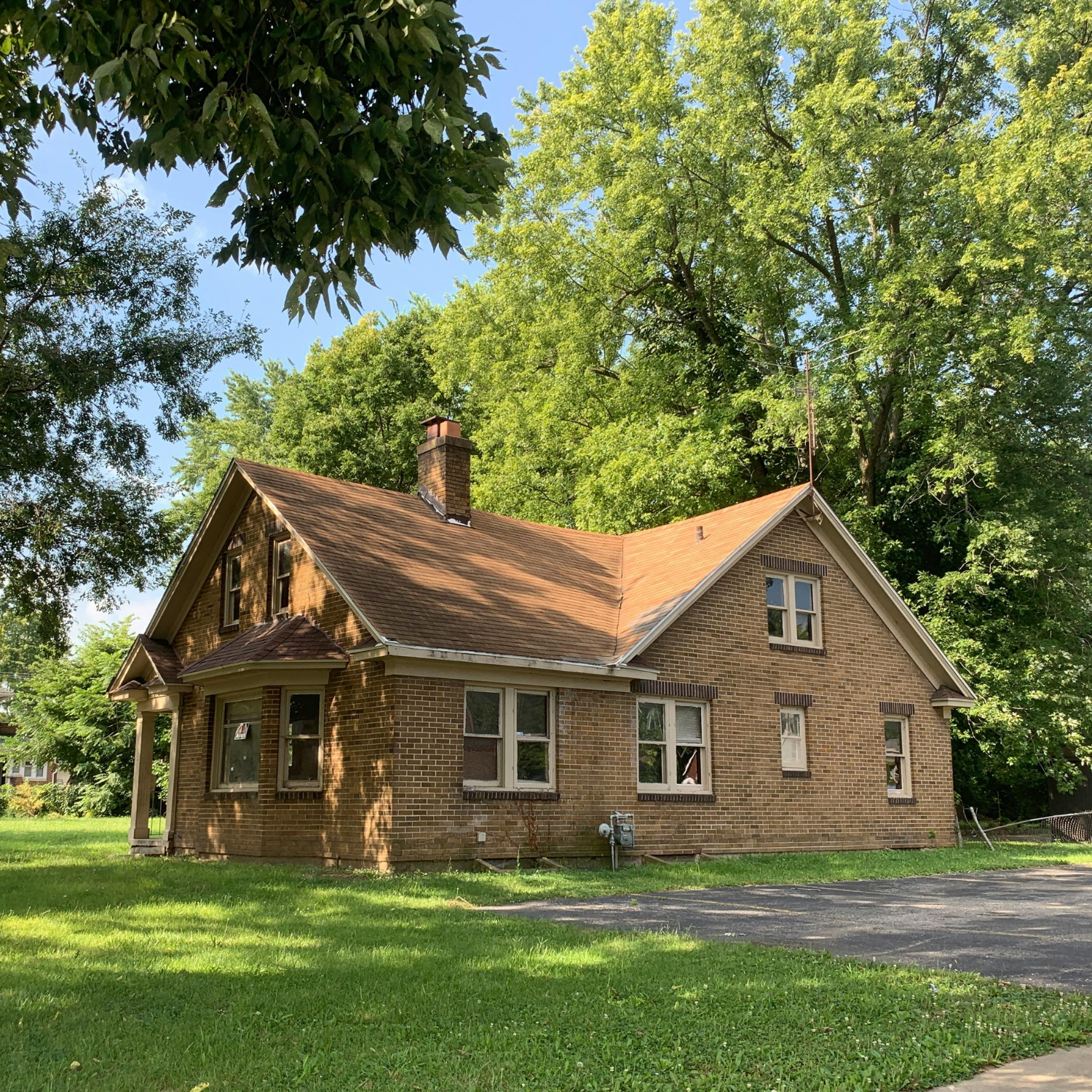 Small tan brick cottage with a red roof beside a gravel drive, surrounded by green trees and lawn.