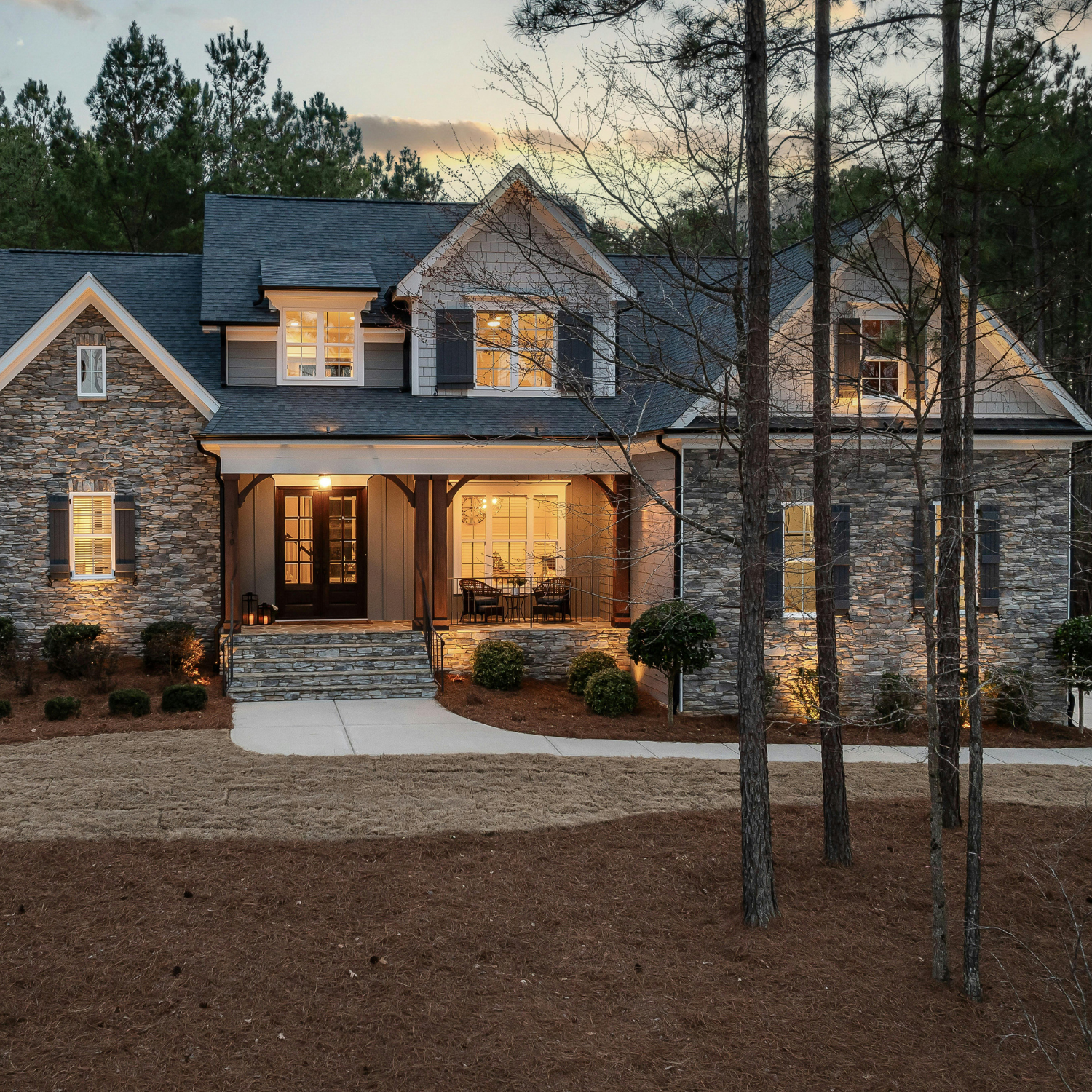 Large two-story stone house at dusk with lit windows, a covered porch, and tall trees in front.