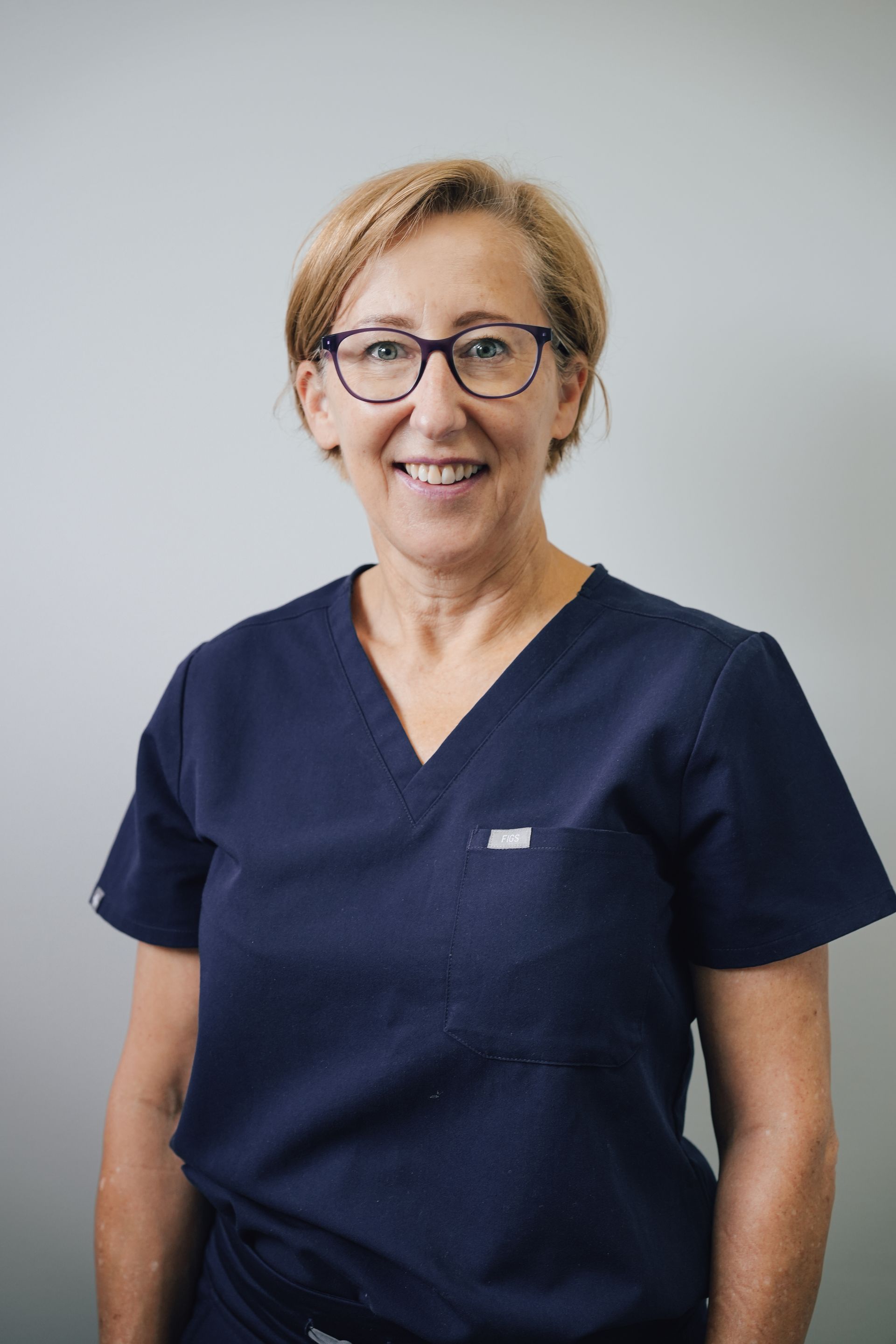 Woman In Navy Blue Scrubs Smiling Against a Light Gray Background — Lord Street Podiatry in Port Macquarie, NSW