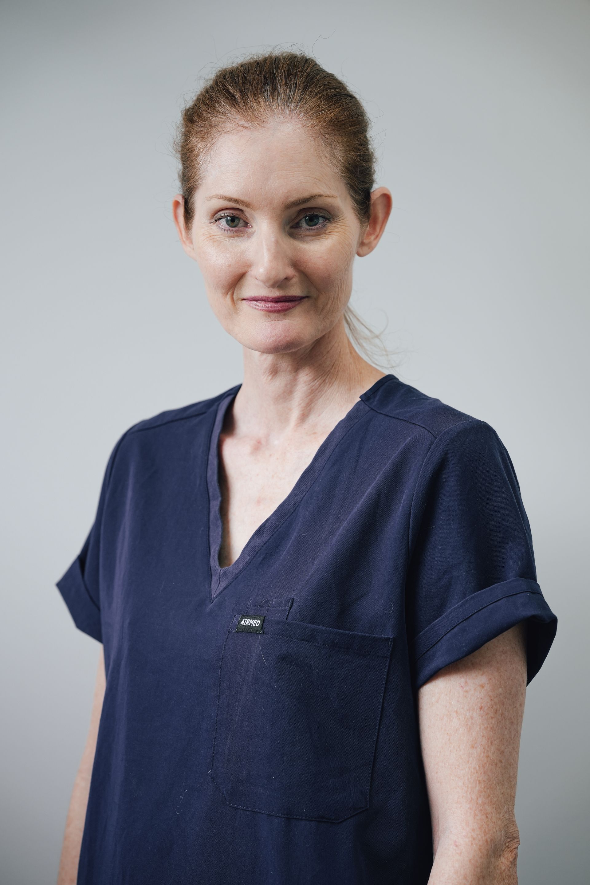 Person In Navy Medical Scrubs Standing Against a Gray Background, Facing Forward With a Slight Smile — Lord Street Podiatry in Port Macquarie, NSW