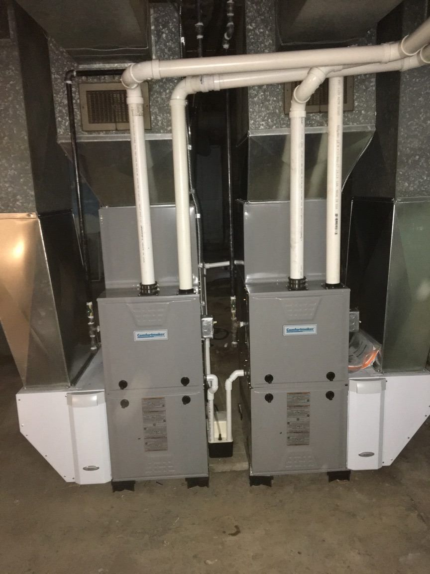 Two HVAC units side-by-side, with white pipes and silver ductwork, in a basement setting.