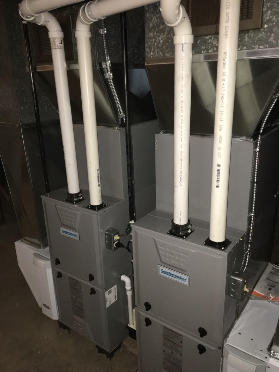 Two gray furnaces with white PVC pipes in a utility room.