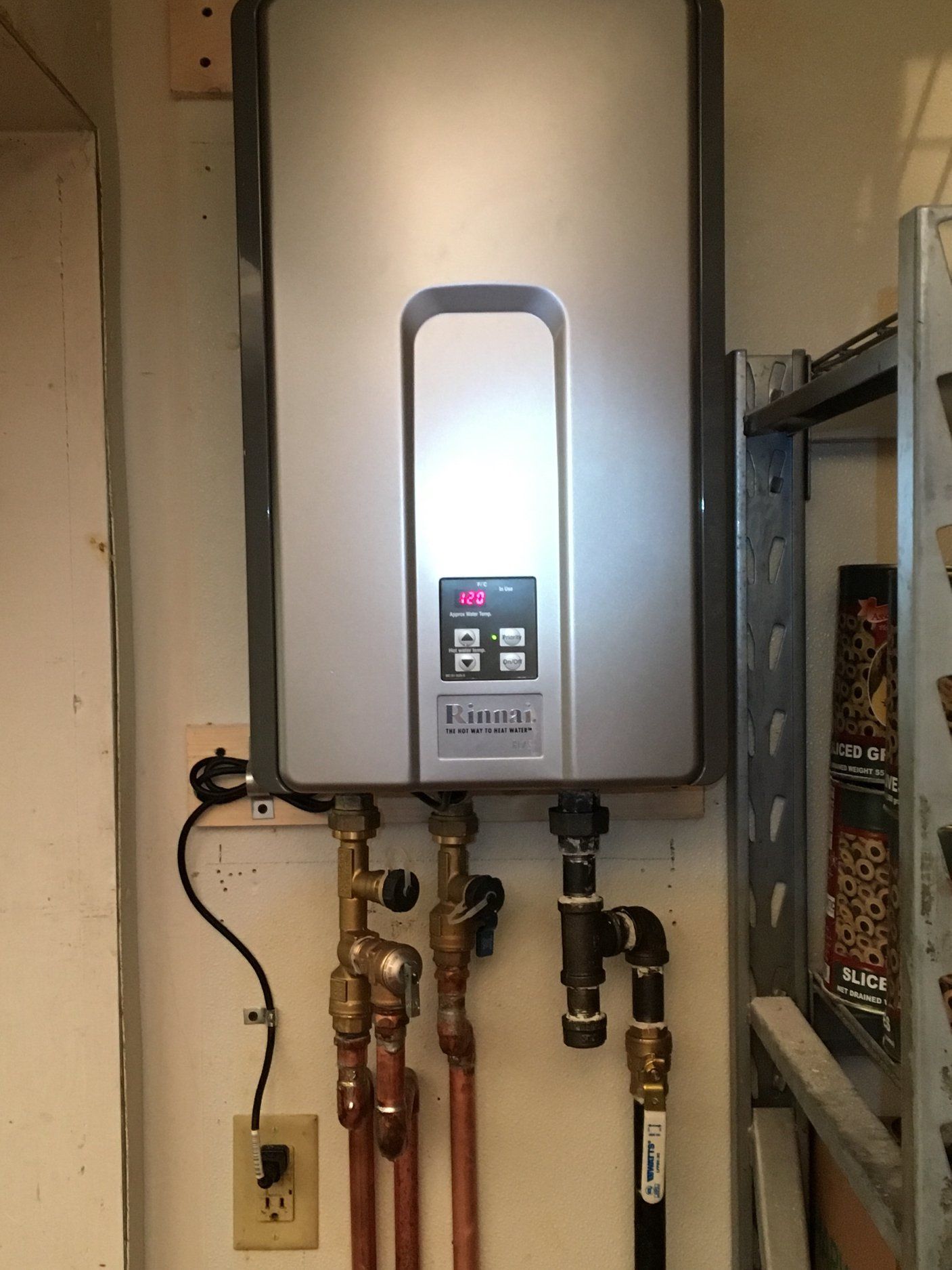 Wall-mounted, silver tankless water heater with copper and black pipes, next to a shelf.