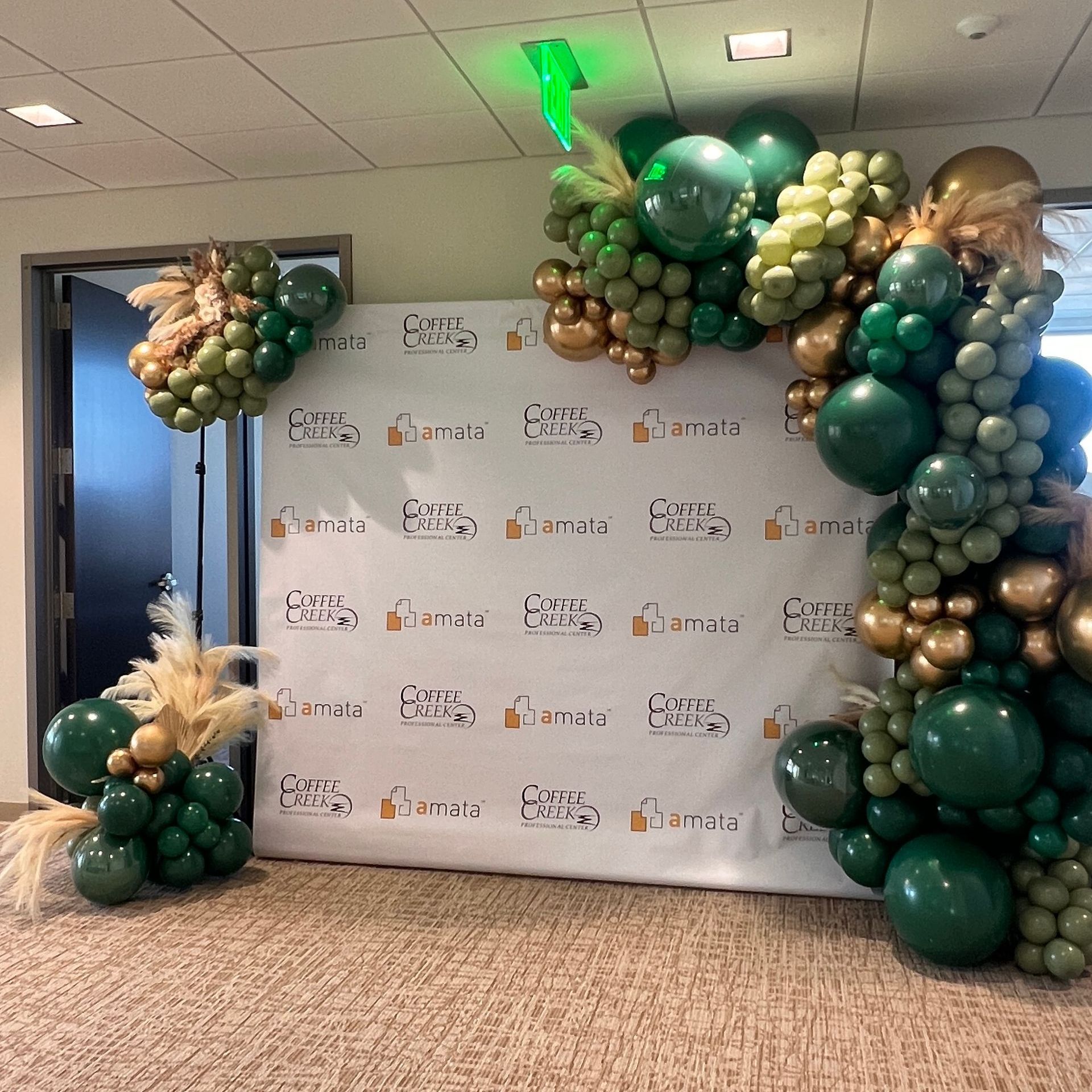 Deluxe balloon garland with green, gold, and sage balloon chains, paired with pampas for an elegant decor and a branded step-and-repeat for a corporate new location in Chesterton, IN.