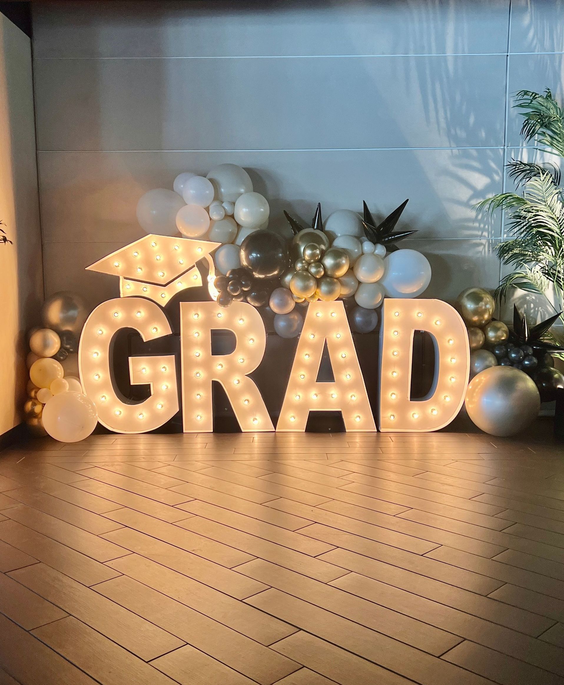 LED marquee letters spelling 'GRAD' with an LED graduation cap topper, complemented by an organic balloon garland in white, black, and gold with black taper balloon accents. Perfect for graduation celebrations and photo opportunities.