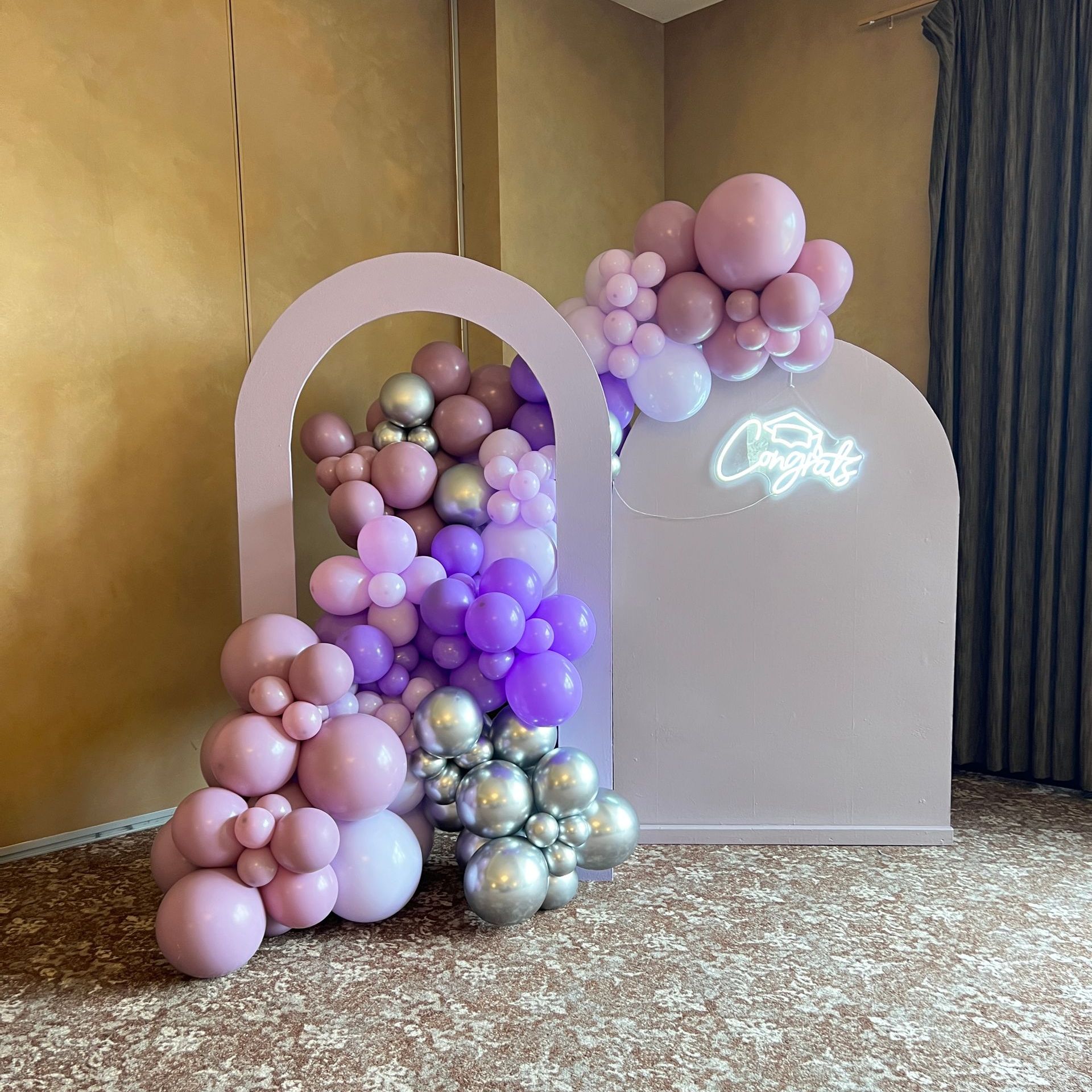 LED marquee letters spelling 'GRAD' with an LED graduation cap topper, complemented by an organic balloon garland in white, black, and gold with black taper balloon accents. Perfect for graduation celebrations and photo opportunities.