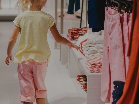 Bambino con maglietta gialla e pantaloni rosa che allunga la mano verso i vestiti in un negozio.