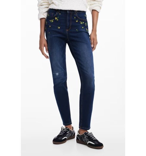 Donna in jeans blu scuro con ricami gialli e scarpe da ginnastica nere.