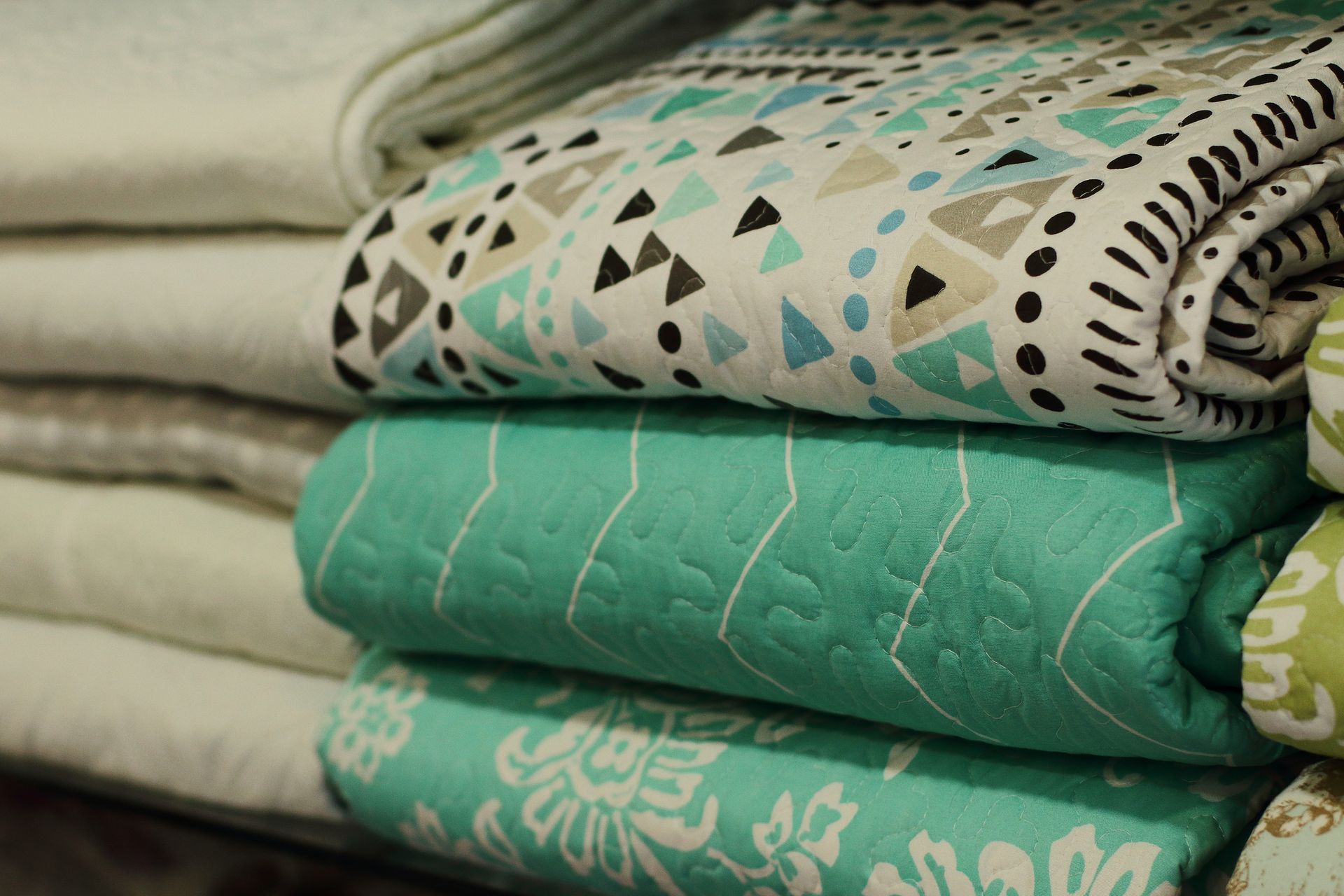 Pila di coperte piegate in vari modelli e colori: verde acqua, bianco e motivi geometrici.