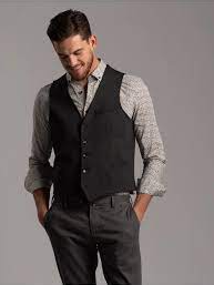 Uomo con gilet nero, camicia fantasia, pantaloni grigi, mani in tasca, sorridente.