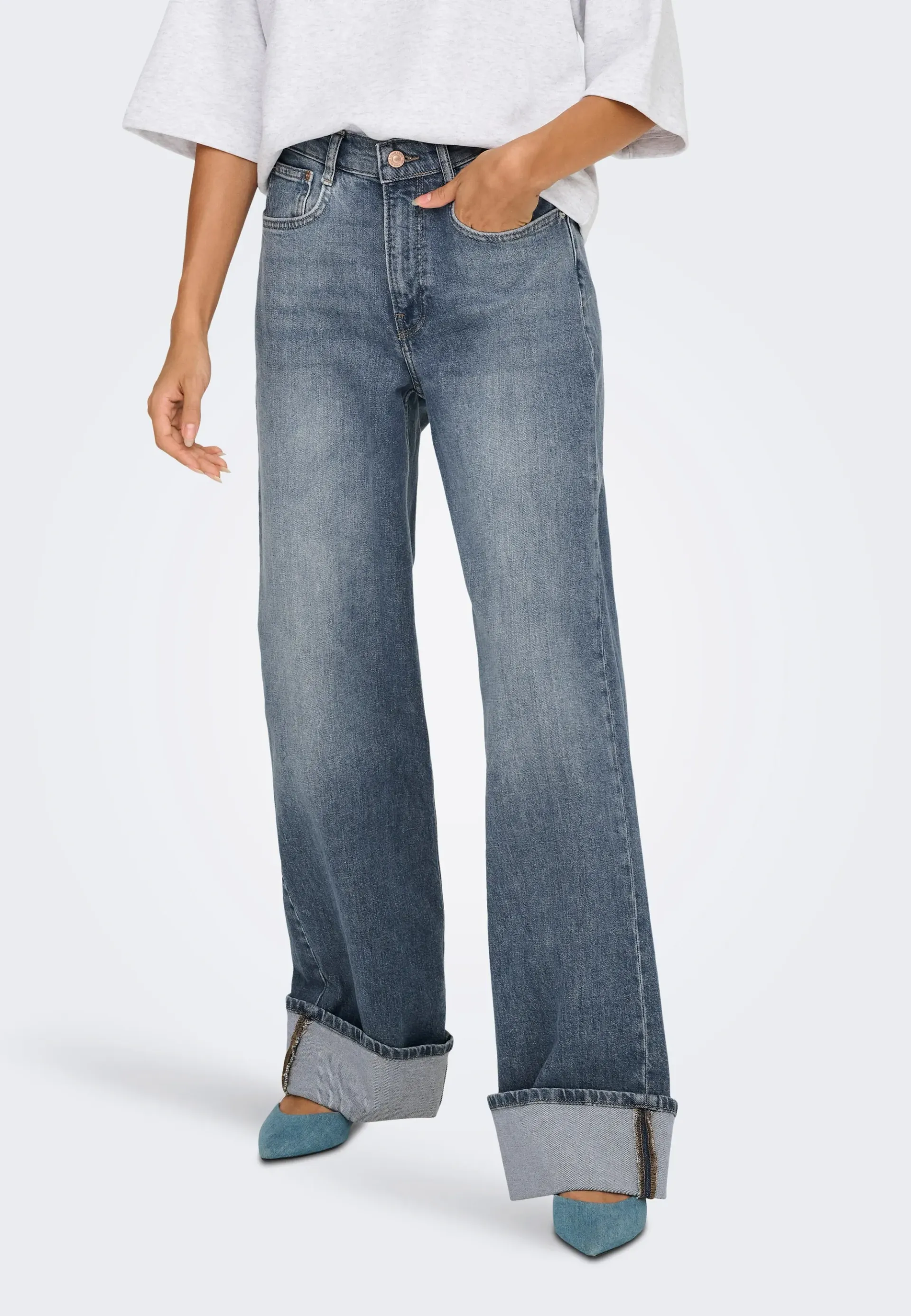 Donna che indossa jeans larghi con lavaggio acido, risvolti e tacchi blu, mani in tasca.