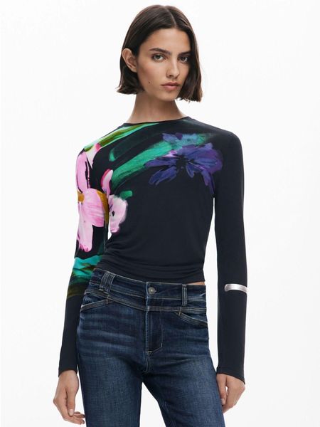 Donna che indossa un top floreale scuro e jeans. È in piedi e guarda avanti.