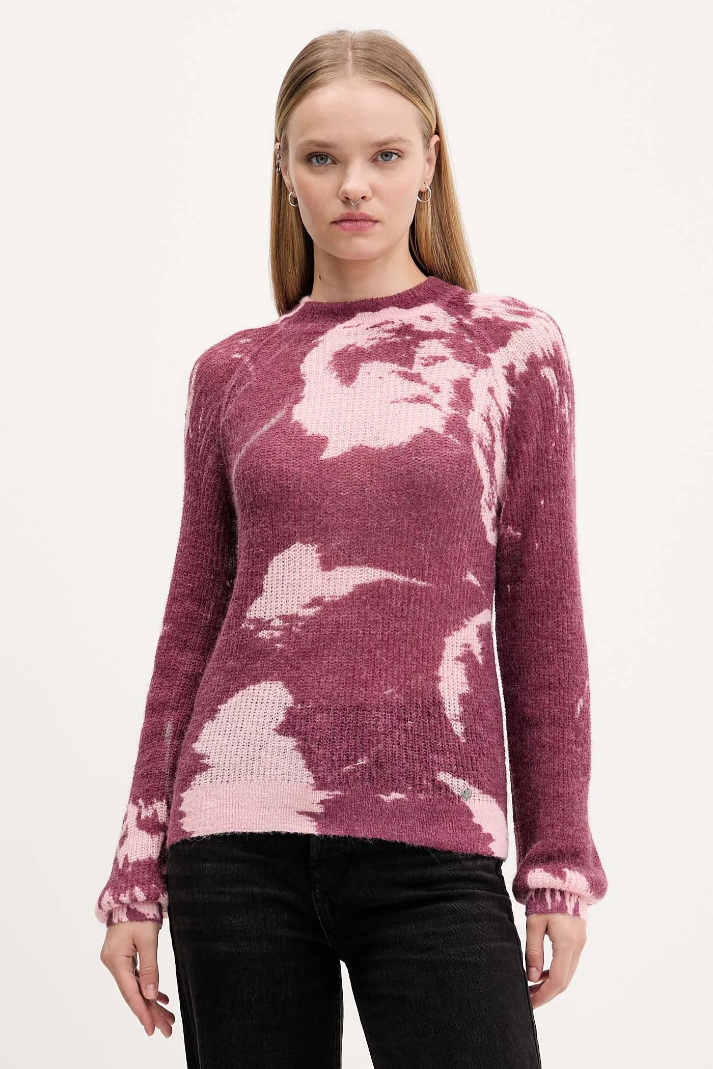 Donna che indossa un maglione rosa e bordeaux, pantaloni neri. Sfondo semplice.